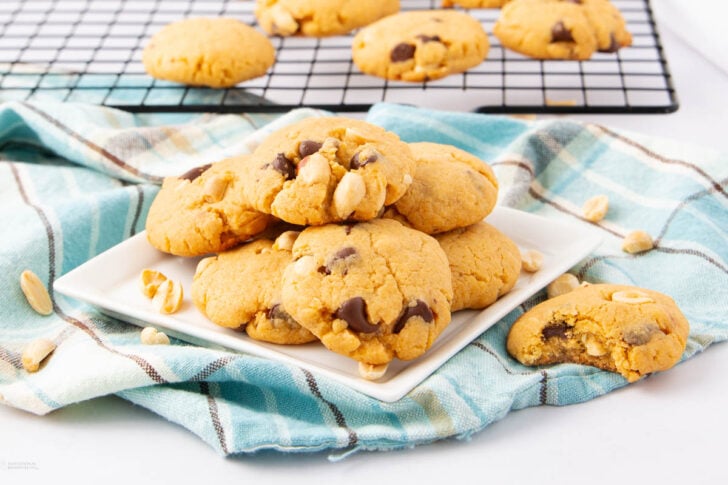A plate of chocolate chip cookies with peanuts sits on a blue plaid cloth. Additional cookies and peanuts are scattered nearby, with more cookies cooling on a wire rack in the background.