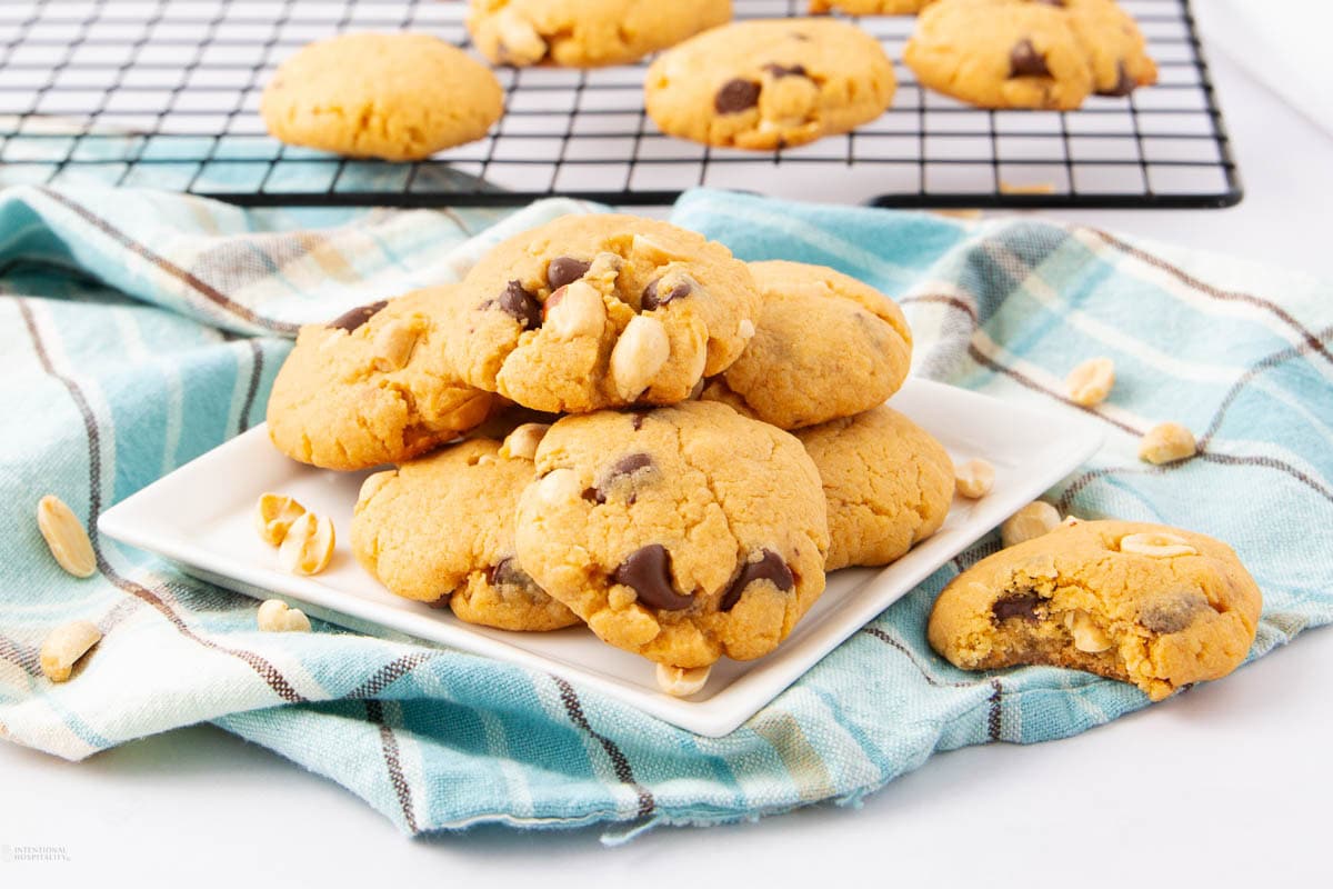 A plate of chocolate chip cookies with peanuts sits on a blue plaid cloth. Additional cookies and peanuts are scattered nearby, with more cookies cooling on a wire rack in the background.
