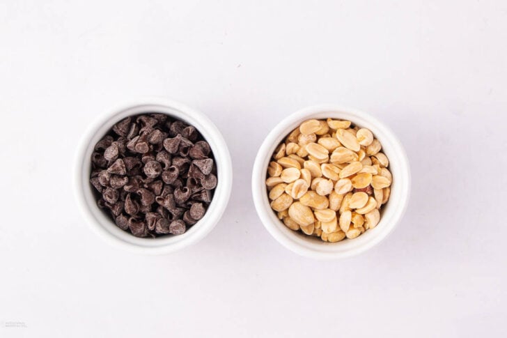 Two small white bowls on a white surface; one bowl contains chocolate chips, and the other contains roasted peanuts.