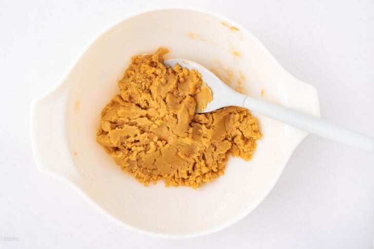 A white mixing bowl containing a mound of light brown cookie dough with a white spatula resting inside, set on a plain white surface.