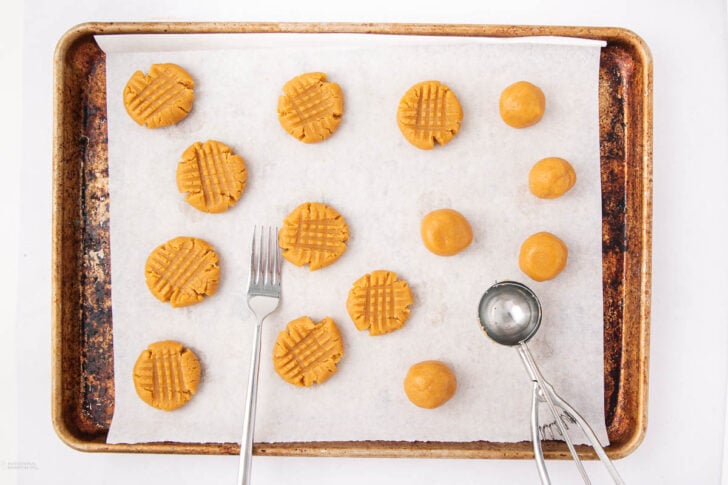 A baking sheet lined with parchment paper holds round balls of peanut butter cookie dough, some pressed with a fork in a crisscross pattern. A metal scoop and fork rest on the tray.