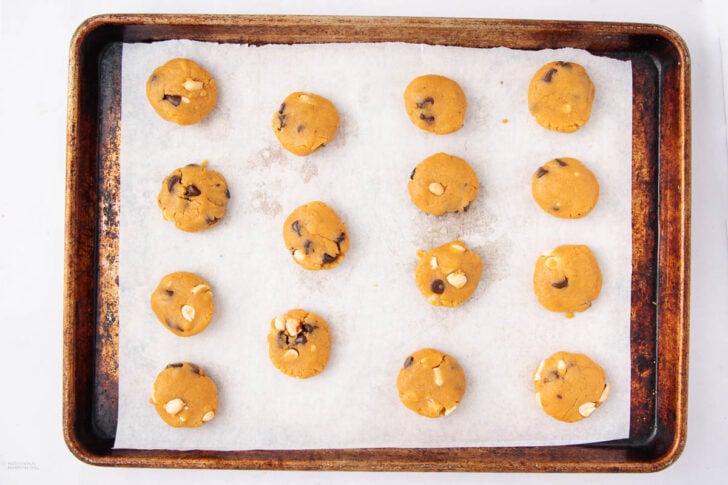 A baking tray lined with parchment paper holds fifteen unbaked, round peanut butter chocolate chip cookie dough balls, spaced evenly apart and ready to be baked.