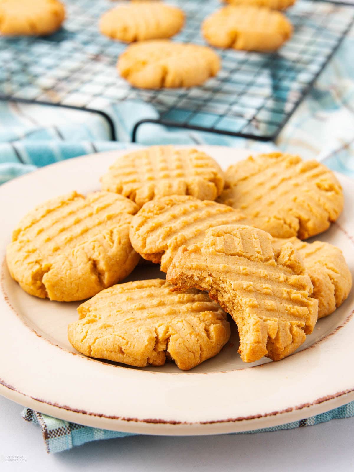 A plate of peanut butter cookies with fork marks on top, one cookie showing a bite taken out. More cookies cooling on a wire rack in the background.