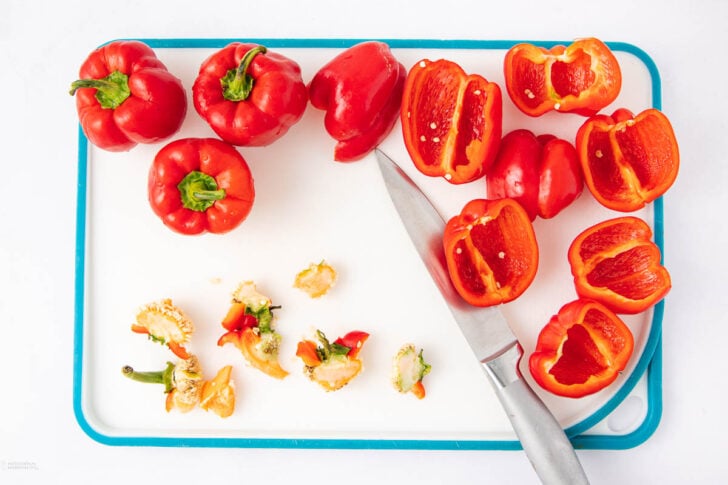 A cutting board with whole and halved red bell peppers, seeds and stems, and a knife on a white background.
