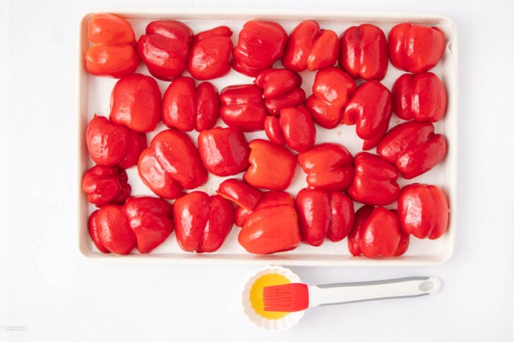 A tray filled with halved red bell peppers, placed cut side down. Next to the tray is a small bowl of oil with a red silicone brush resting on top. The background is white.