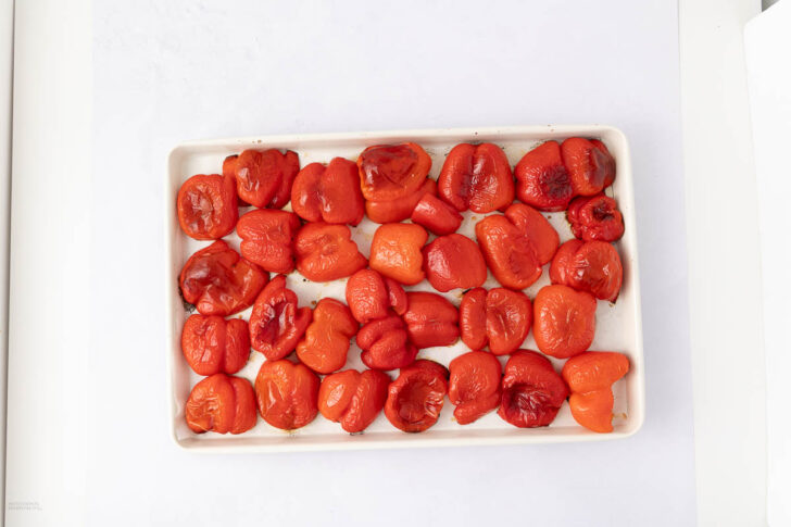 A white rectangular tray filled with roasted red bell peppers, arranged in rows on a white surface. The peppers are slightly charred and have a shiny, softened appearance.
