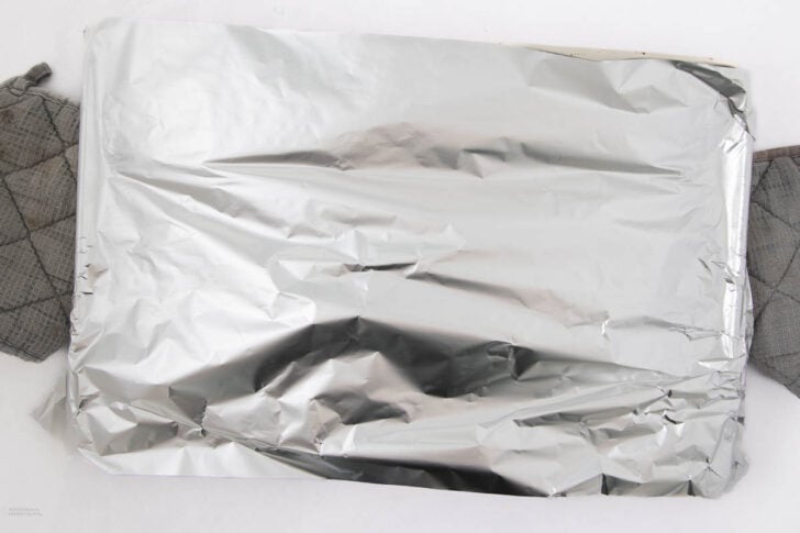 A rectangular baking tray covered with crumpled aluminum foil sits on two gray oven mitts against a white background.