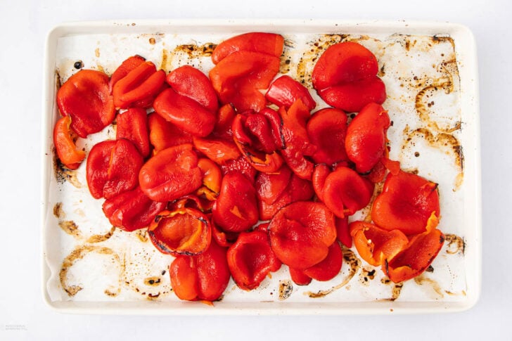 Red roasted bell pepper pieces are spread out on a white baking tray lined with stained parchment paper, showing charred spots and juices from roasting.