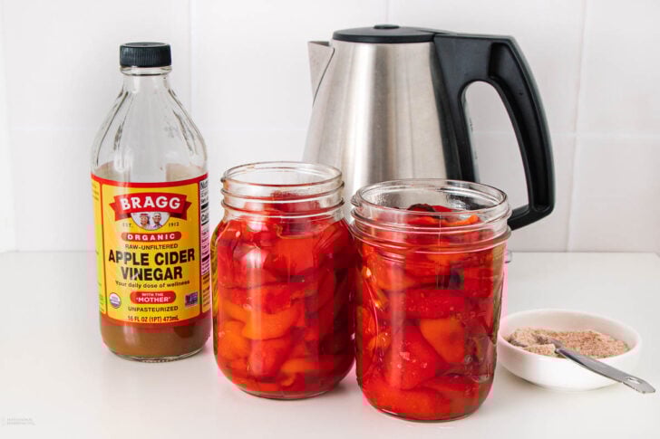 Two jars filled with red bell peppers in liquid sit on a counter next to a bottle of apple cider vinegar, a bowl of salt, and an electric kettle against a white tiled background.