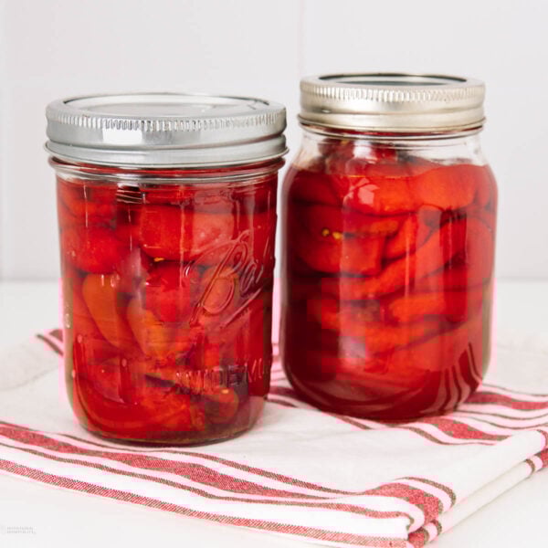 Two glass jars filled with red pickled peppers, sealed with metal lids, sit on a folded white and red striped cloth against a white background.