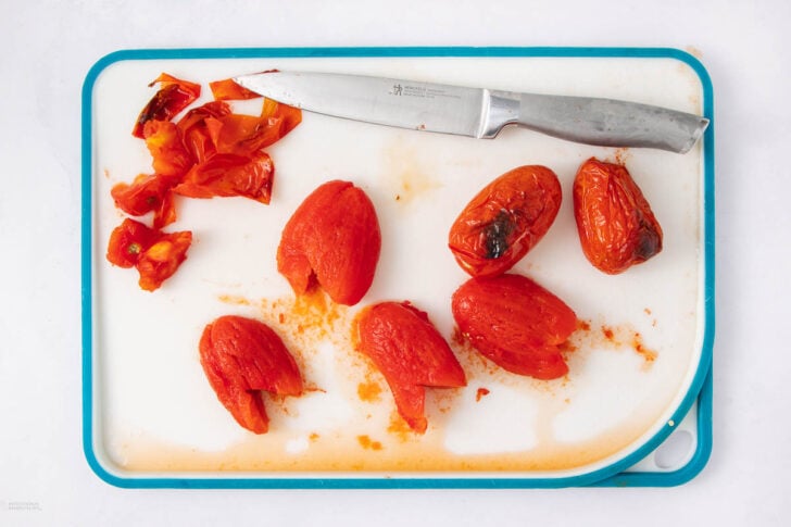 A cutting board with a silver knife, several peeled tomatoes, and tomato skins arranged on it. The surface is white with blue edges, and there are tomato juices visible on the board.