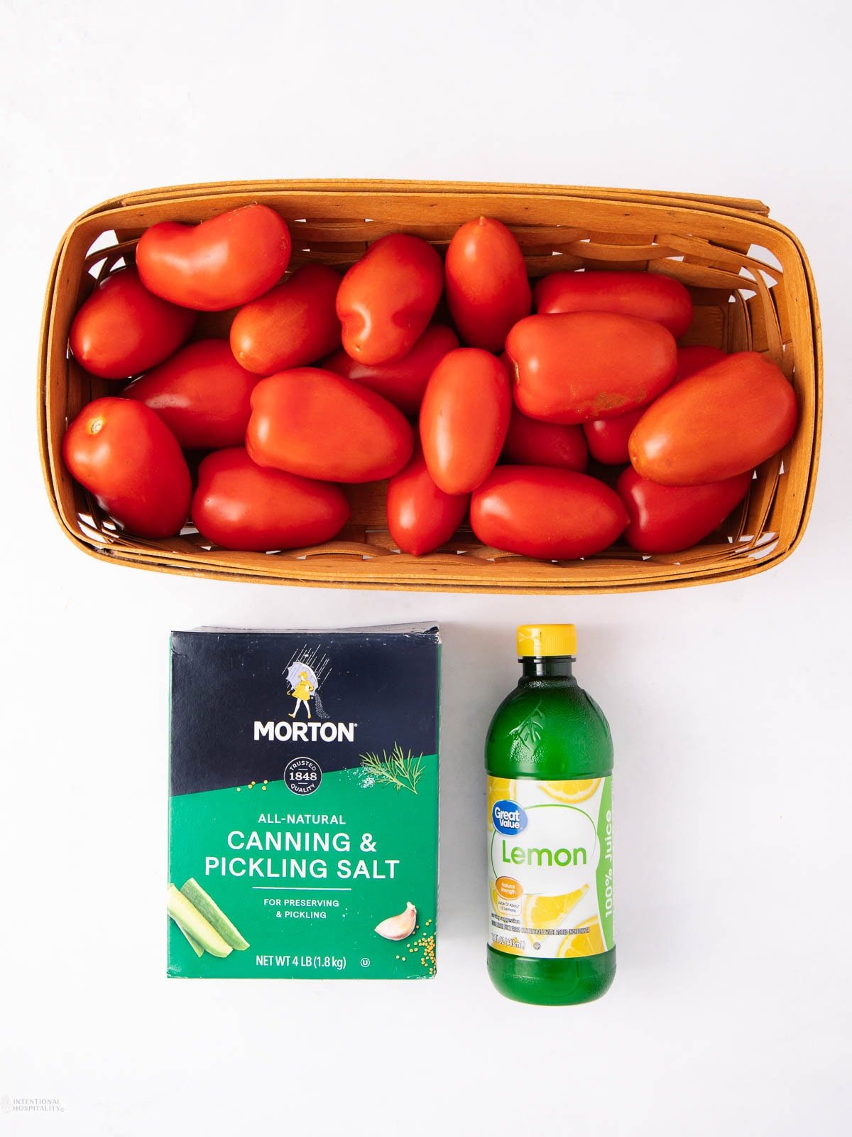 A basket of roma tomatoes sits above a box of canning and pickling salt and a bottle of lemon juice, all arranged on a white surface.