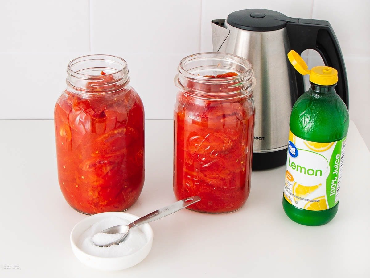 Two glass jars filled with chopped tomatoes sit on a counter next to a bowl of salt with a measuring spoon, a bottle of lemon juice, and a stainless steel electric kettle in the background.