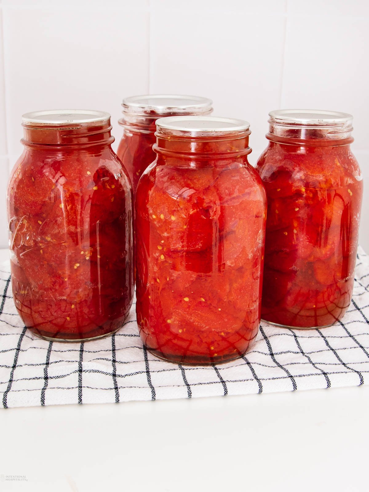 Four glass jars filled with canned tomatoes sit on a black and white checkered towel, placed on a white countertop with a tiled white background.