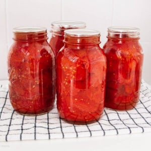 Four glass jars filled with preserved tomatoes in liquid are placed on a white and black checkered cloth, against a white tiled background.