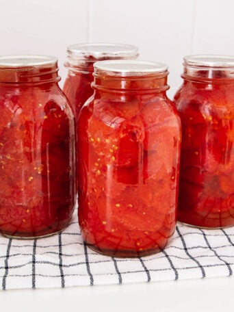 Four glass jars filled with preserved tomatoes in liquid are placed on a white and black checkered cloth, against a white tiled background.