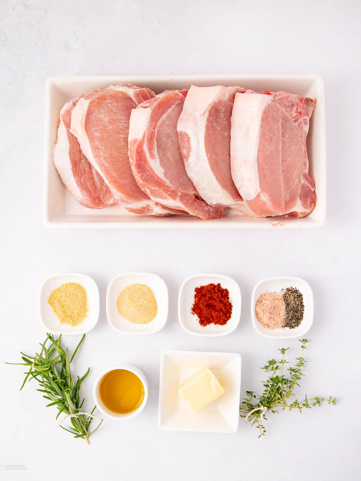 Raw pork chops are arranged in a white rectangular dish. Below, small bowls contain garlic powder, paprika, salt and pepper, olive oil, and butter. Fresh rosemary and thyme sprigs are placed on the side.