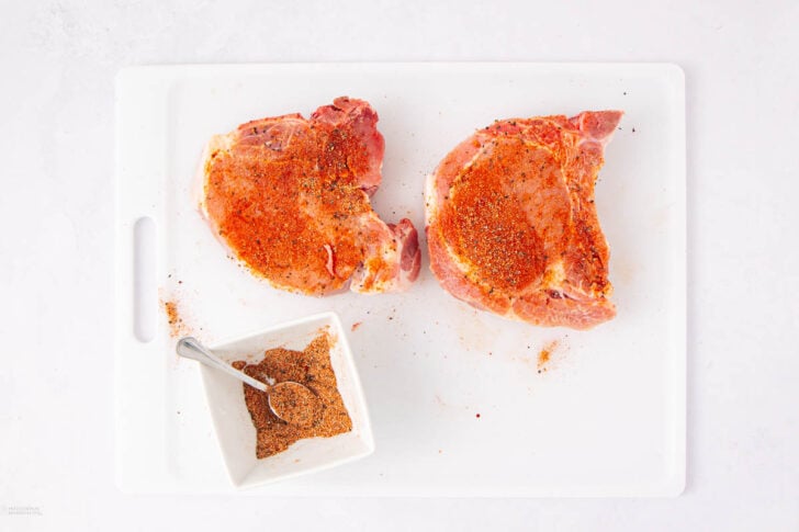 Two raw pork chops coated with spice rub on a white cutting board, next to a small white dish filled with more seasoning and a spoon.