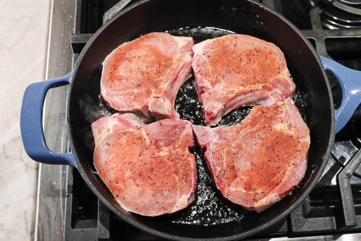 Four seasoned pork chops are sizzling in a blue-handled cast iron skillet on a stovetop. The pork chops are partially overlapping and appear to be freshly placed in the pan for cooking.