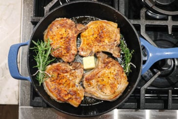 Four seasoned pork chops are sizzling in a cast iron skillet with a pat of butter and sprigs of fresh rosemary, on a stovetop with black grates and a blue handle on the pan.