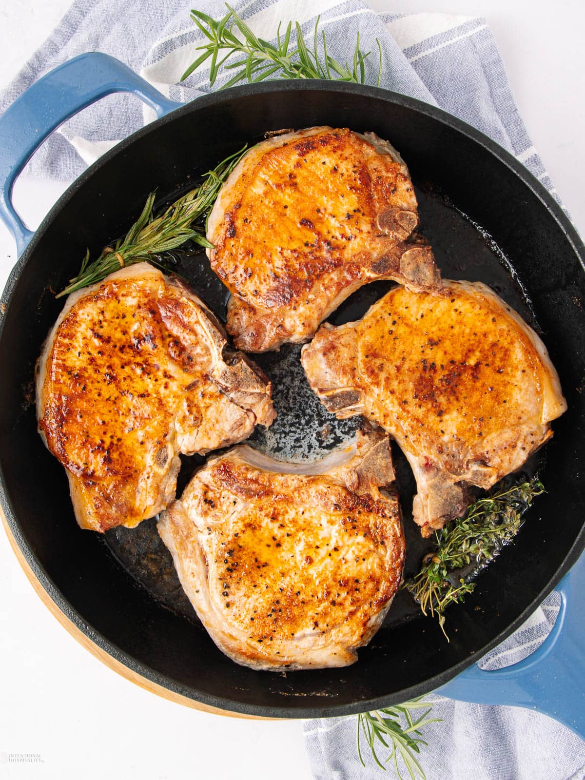 Four golden-brown pork chops are sizzling in a black skillet with sprigs of fresh rosemary, set on a blue and white striped towel. The pan has a blue handle and the chops are evenly spaced, each with a crispy, seared surface.