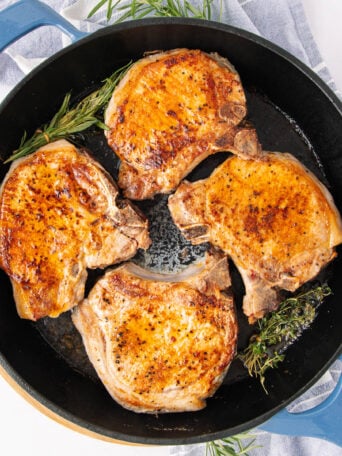 Four golden-brown pork chops are cooking in a black skillet with sprigs of fresh rosemary and thyme, placed on a striped kitchen towel. The meat appears juicy and well-seasoned.