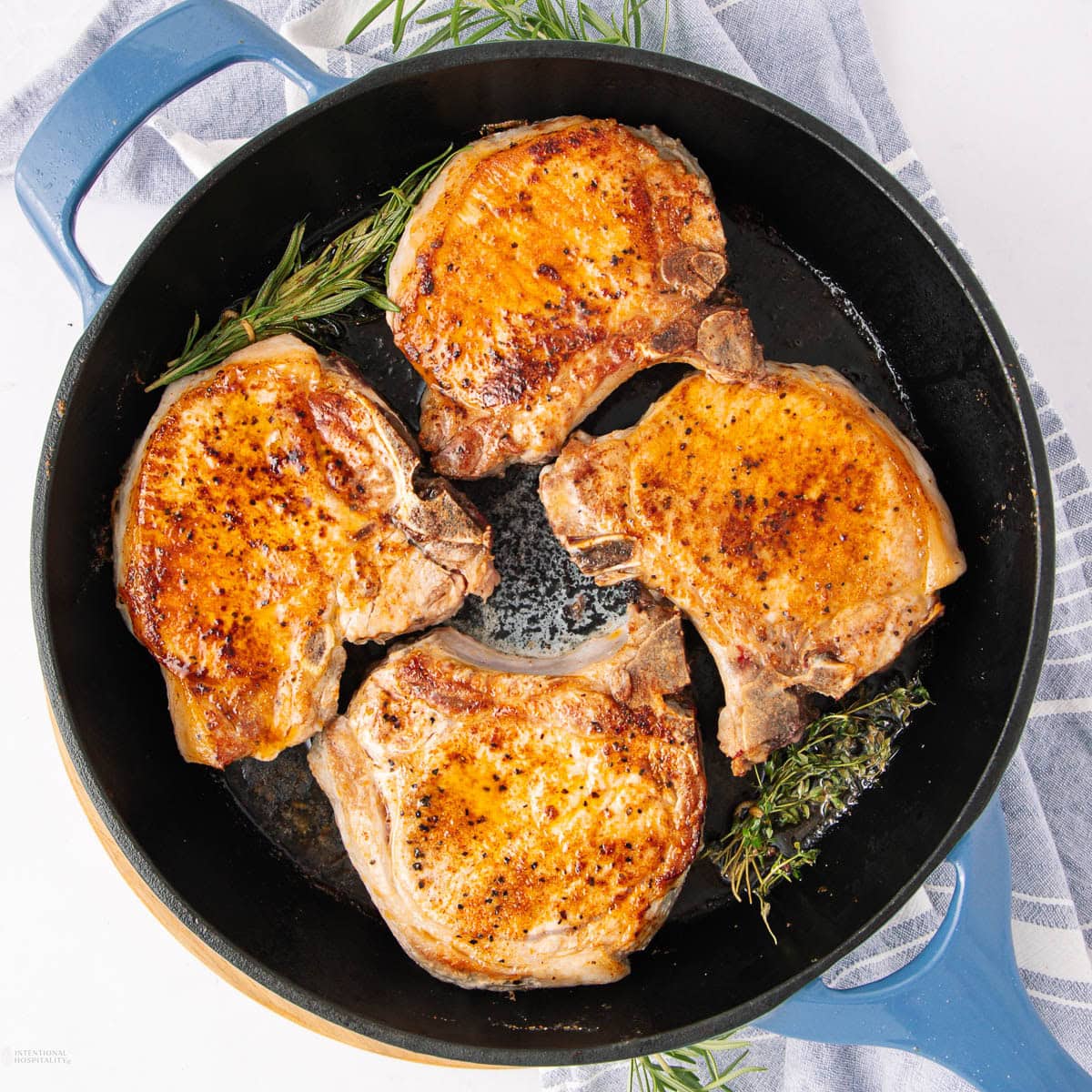 Four golden-brown pork chops are cooking in a black skillet with sprigs of fresh rosemary and thyme, placed on a striped kitchen towel. The meat appears juicy and well-seasoned.