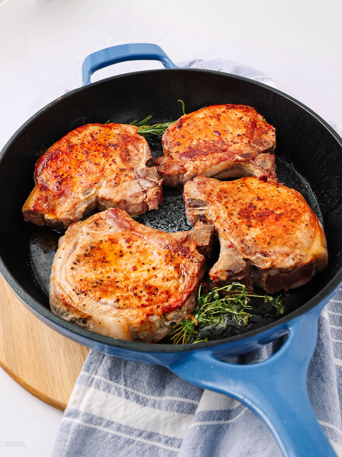 A blue skillet with four seasoned pork chops cooking on a bed of fresh herbs, resting on a wooden board and a striped kitchen towel.