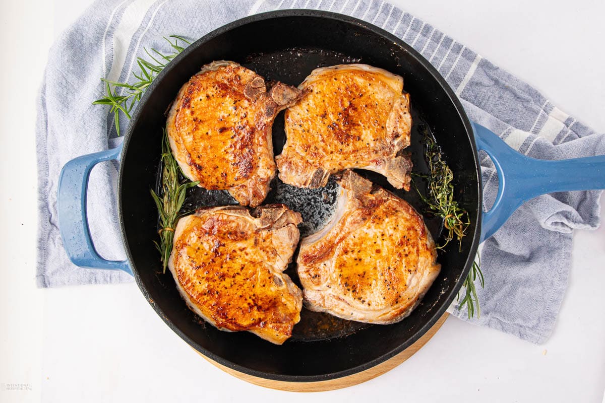 A black skillet with four golden-brown, cooked pork chops, garnished with fresh rosemary and thyme, sits on a blue and white striped towel.