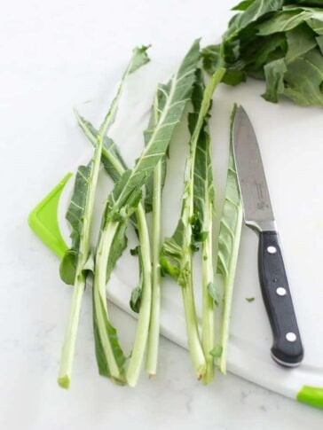 A cutting board with collard green stems, a paring knife, and leafy greens in the background—perfect for prepping ingredients for your favorite canned cowboy candy recipe. The stems are separated from the leaves and arranged beside the knife.