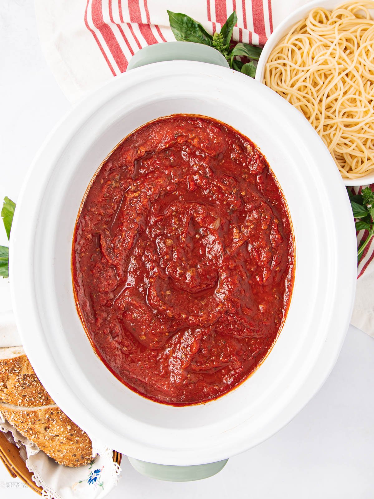 A white slow cooker filled with red tomato pasta sauce sits next to a bowl of cooked spaghetti, a red-striped towel, and a plate of bread slices on a white surface.
