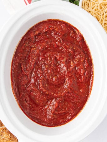 A white bowl filled with thick, rich red marinara sauce sits next to a plate of plain spaghetti noodles and a slice of multigrain bread, all arranged on a white surface.