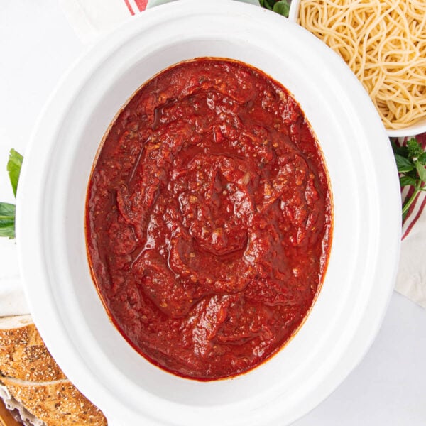 A white bowl filled with thick, rich red marinara sauce sits next to a plate of plain spaghetti noodles and a slice of multigrain bread, all arranged on a white surface.