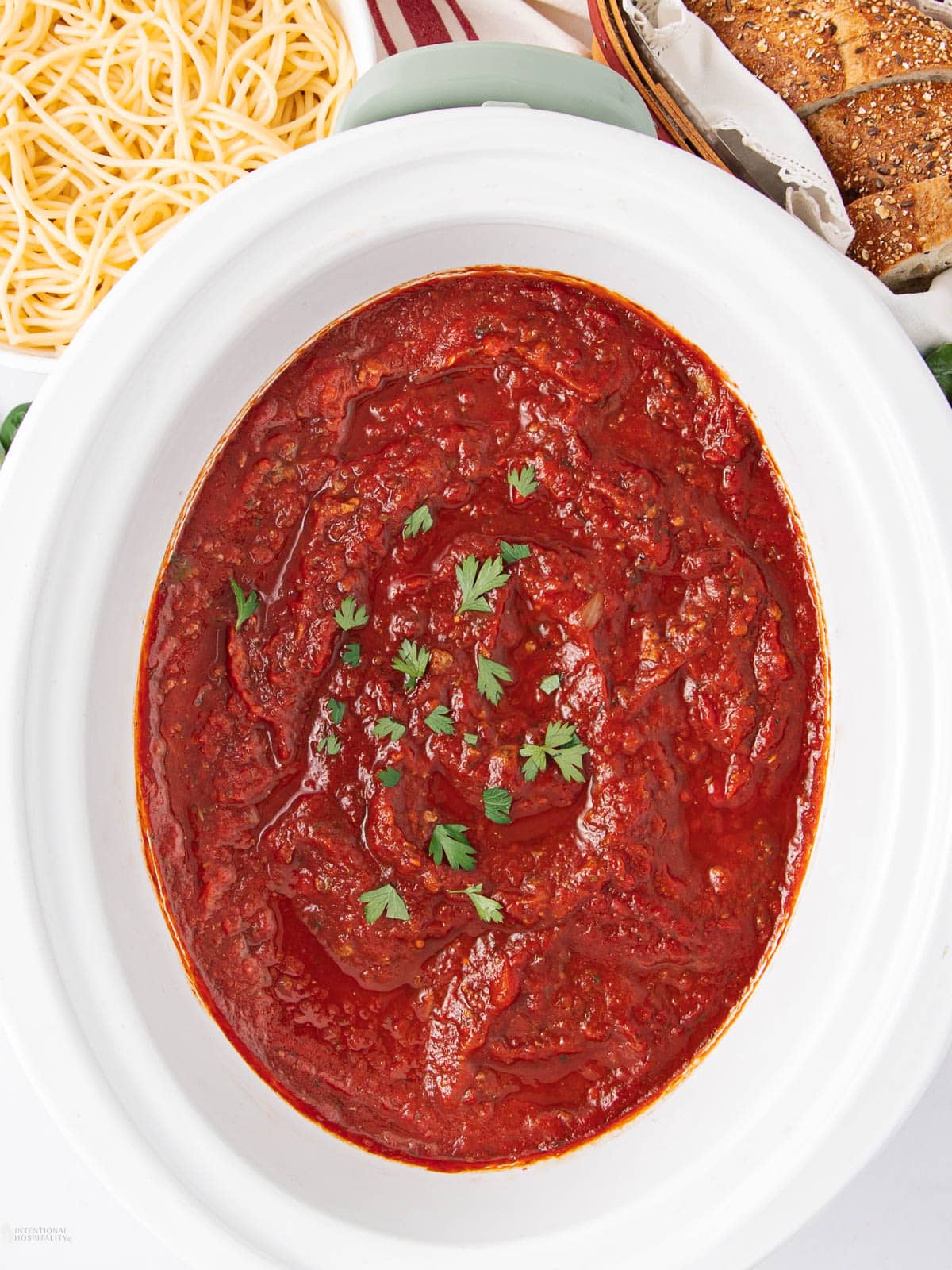 A white slow cooker filled with thick red marinara sauce, garnished with parsley. Nearby are a plate of cooked spaghetti and a basket of sliced bread.
