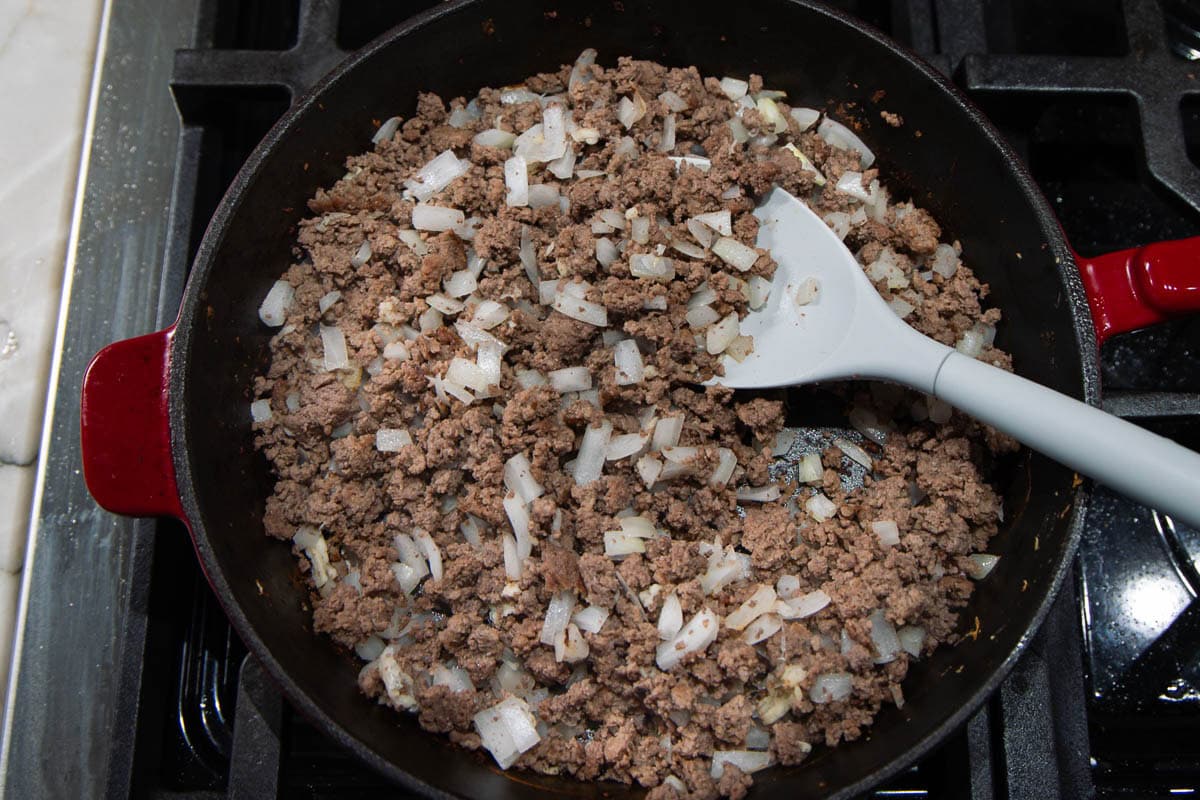 Ground beef and chopped onions cooking in a red skillet on a stovetop, with a gray spatula resting in the pan.
