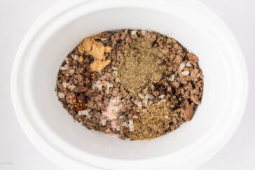 Ground beef and diced onions in a white slow cooker, topped with various seasonings including dried herbs, ground spices, and red pepper flakes, ready to be mixed.