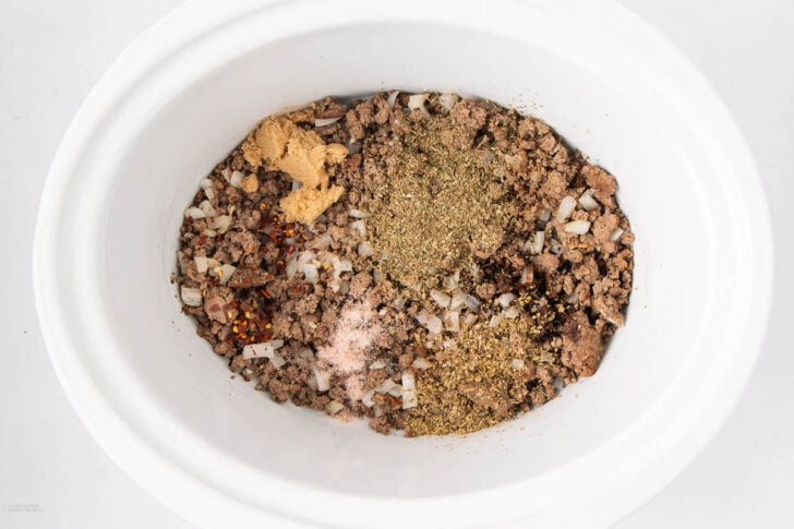 Ground beef and diced onions in a white slow cooker, topped with various seasonings including dried herbs, ground spices, and red pepper flakes, ready to be mixed.