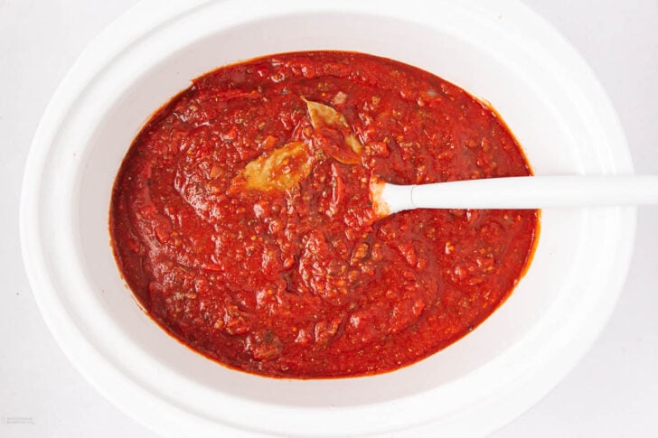 A white slow cooker filled with thick, red tomato sauce containing ground meat and a bay leaf. A white spoon rests inside the sauce.