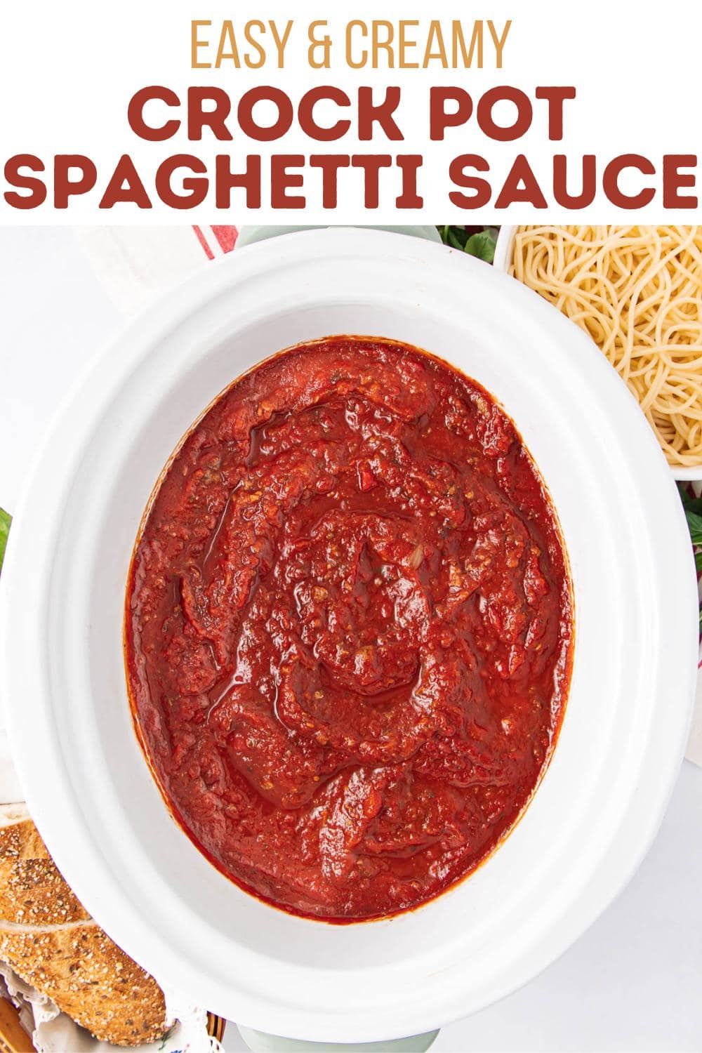 Overhead view of a white crock pot filled with red spaghetti sauce, surrounded by a plate of cooked spaghetti and a plate of bread. Text above reads "Easy & Creamy Crock Pot Spaghetti Sauce.