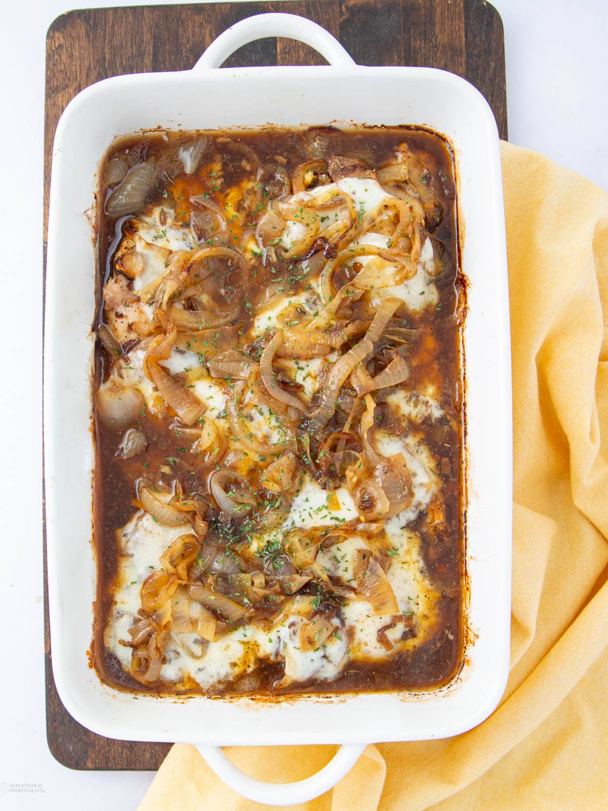 A white baking dish filled with baked chicken topped with caramelized onions and melted cheese in a rich brown sauce, set on a wooden board with a yellow cloth beside it.
