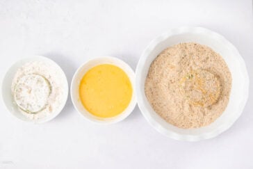 Three bowls arranged left to right: the first with flour and a coated slice, the second with beaten eggs, and the third with breadcrumbs and a slice being coated.