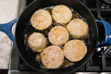 Seven breaded slices of green tomato are frying in oil in a black skillet on a stovetop. The tomatoes are golden brown around the edges, with some bubbling oil visible.