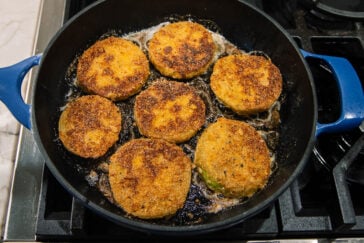 Seven slices of breaded and fried green tomatoes are sizzling in a black skillet with a blue handle on a stovetop. Bubbles from frying oil surround the golden-brown tomato slices.