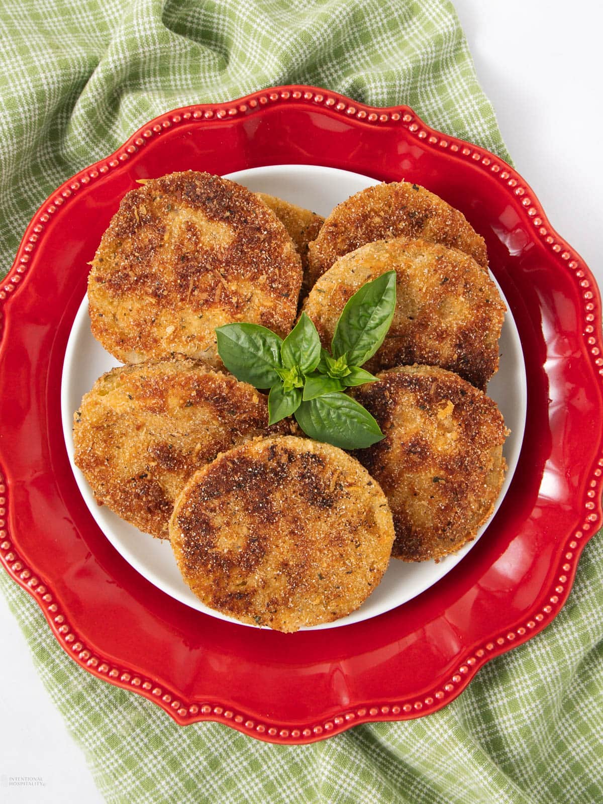 A white plate on a red decorative dish holds several golden-brown fried green tomato slices, garnished with fresh basil leaves, atop a green plaid cloth.