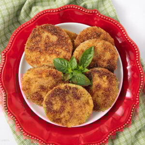 A plate of six golden-brown fried green tomato slices garnished with fresh basil, arranged on a white plate with a decorative red border on top of a green checkered cloth.