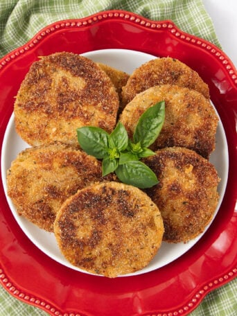A plate of six golden-brown fried green tomato slices garnished with fresh basil, arranged on a white plate with a decorative red border on top of a green checkered cloth.