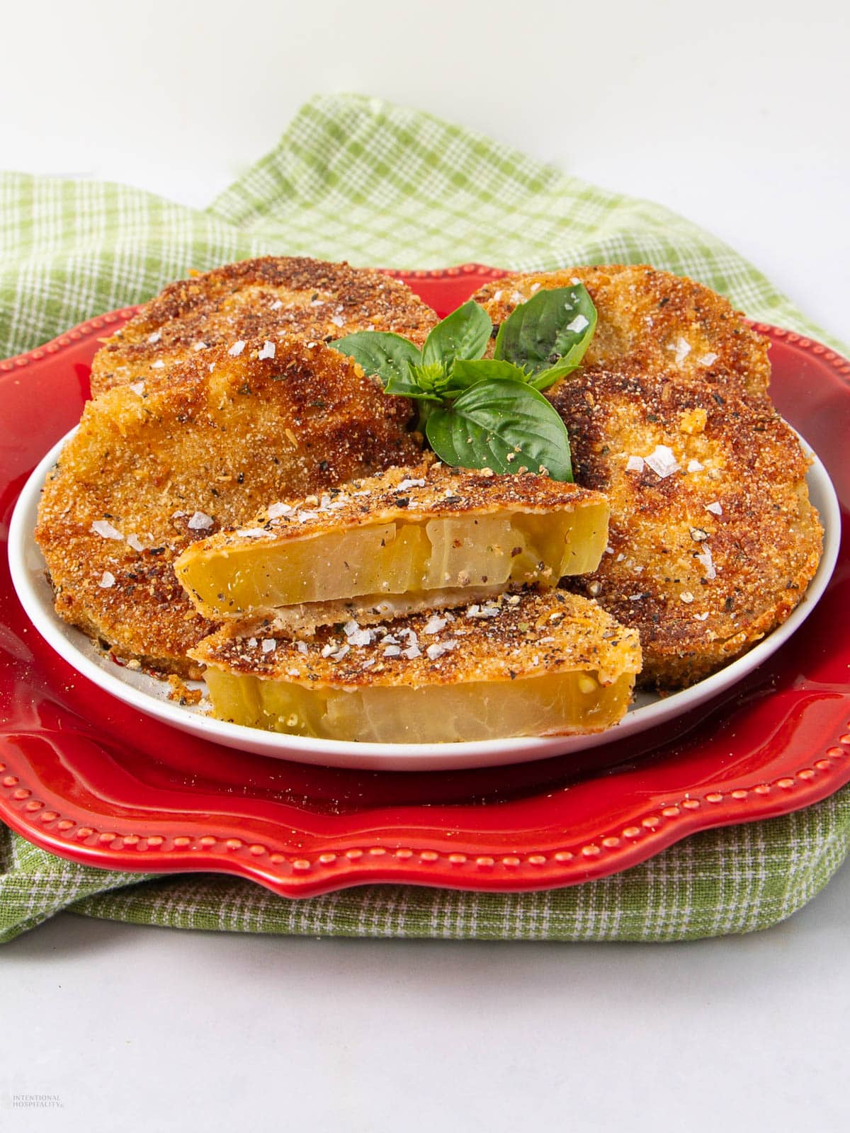 A plate of crispy, golden-brown fried green tomatoes is served on a red dish, garnished with fresh basil. One tomato is sliced to show its juicy yellow-green interior. A green checkered cloth lies underneath.