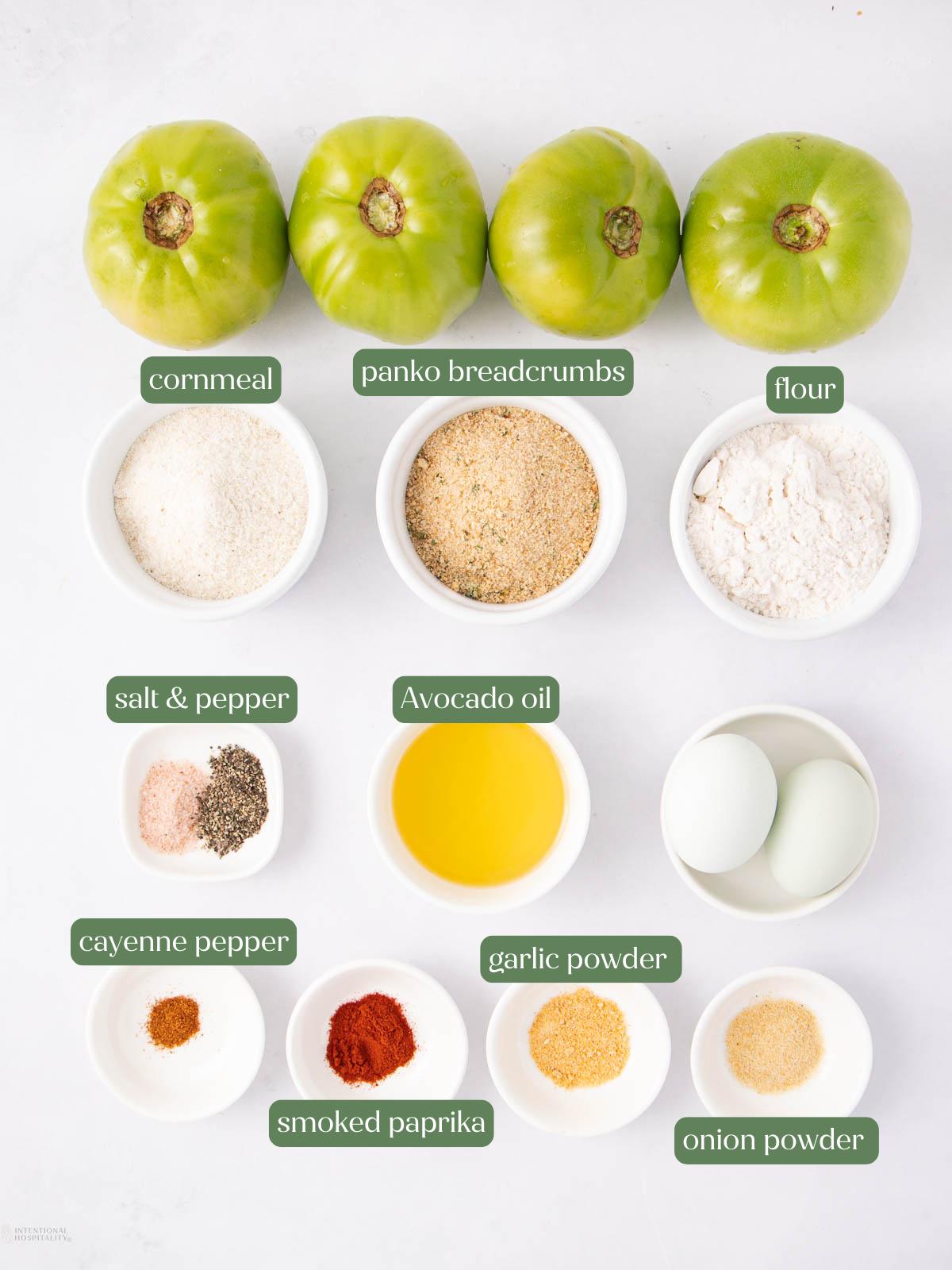 Overhead view of ingredients for fried green tomatoes: three green tomatoes, cornmeal, panko breadcrumbs, flour, salt & pepper, avocado oil, eggs, cayenne pepper, smoked paprika, garlic powder, and onion powder.
