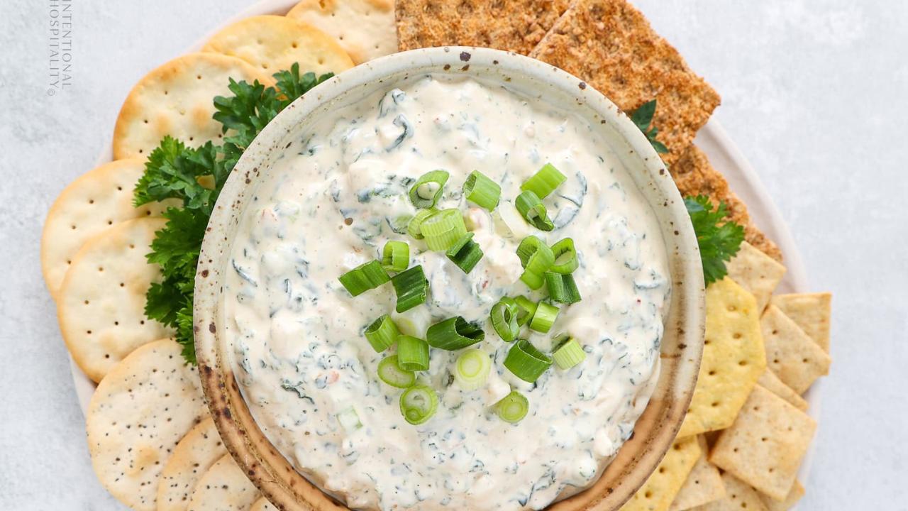 A bowl of creamy spinach dip topped with chopped green onions, surrounded by assorted crackers and fresh parsley on a white plate.