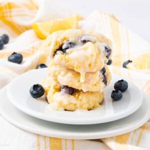 A stack of three glazed blueberry lemon cookies sits on a white plate, surrounded by fresh blueberries, with a yellow and white checkered cloth and a lemon wedge in the background.